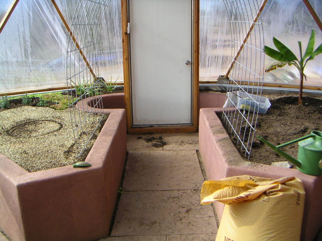 Interior view of a geodesic dome greenhouse showing a neatly arranged stucco raised bed filled with soil ready for planting, with a bag of soil and gardening tools nearby.