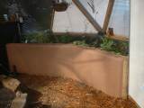 A stucco raised bed inside a greenhouse, freshly built with a smooth finish, ready to be filled with soil and plants, with a scattering of straw on the ground nearby."