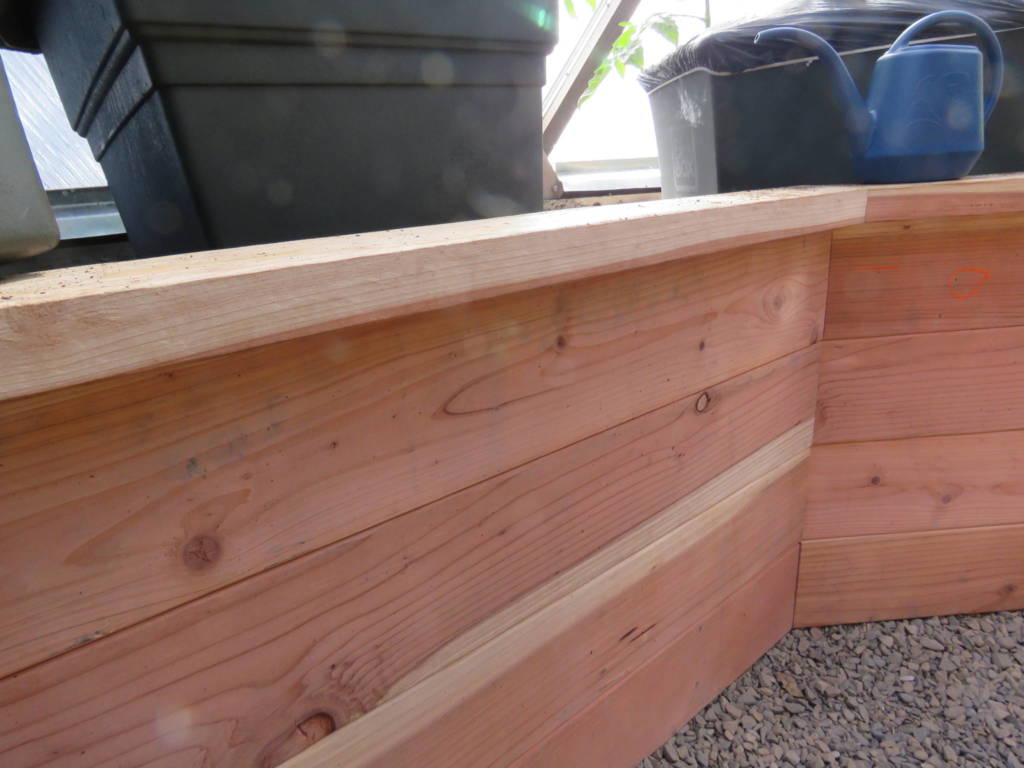Close-up of the smooth top cap and side planks of a wooden raised garden bed, with gardening containers in the background, inside a bright greenhouse.