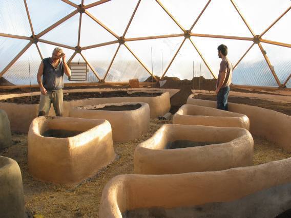Two people contemplating the setup of stucco raised beds inside a spacious geodesic dome, with the beds arranged in a circular pattern on a dirt floor.