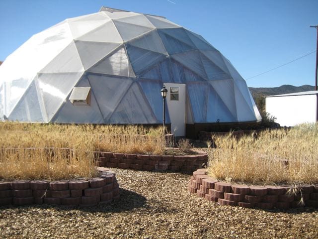 The exterior of a geodesic dome greenhouse flanked by Pavestone raised beds in a semi-circular arrangement, standing ready to nurture plants, against a backdrop of wild grass and clear skies.