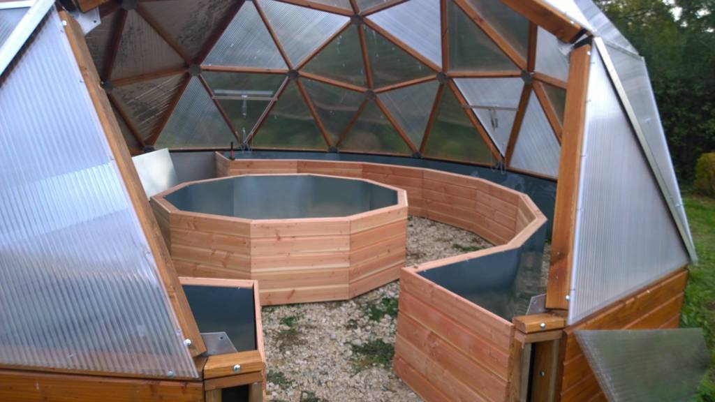 A series of empty curved raised wooden beds inside a geodesic dome greenhouse, with a transparent ceiling reflecting the sky at dusk redwood