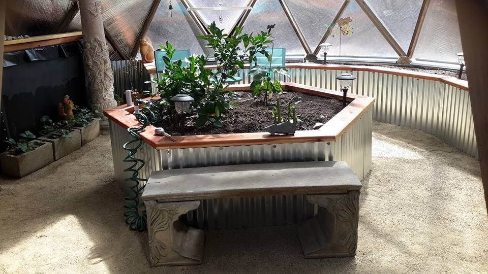 A serene corner inside a greenhouse with a built-in bench, featuring a raised garden bed edged with corrugated aluminum siding, housing healthy plants and providing a peaceful spot for garden contemplation.