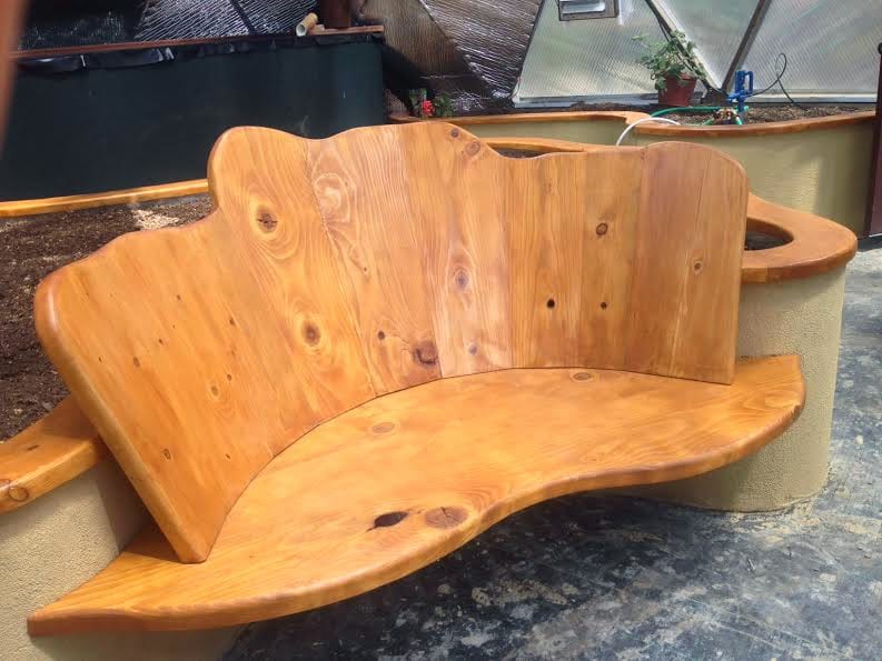 A uniquely designed wooden bench with a curved backrest and seat, crafted from polished wood, creating a warm and inviting rest area within a greenhouse setting.