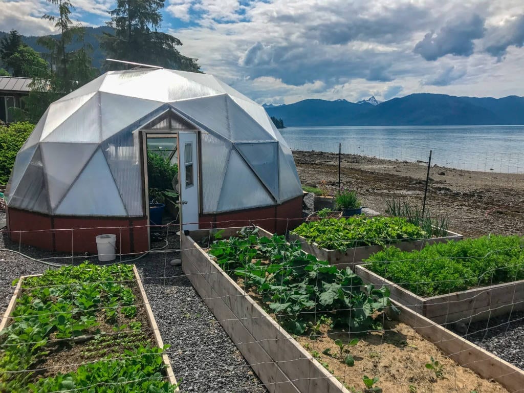 22' Alaska Greenhouse Growing Dome with raised beds filled with plants in the front and water and mountains in the background