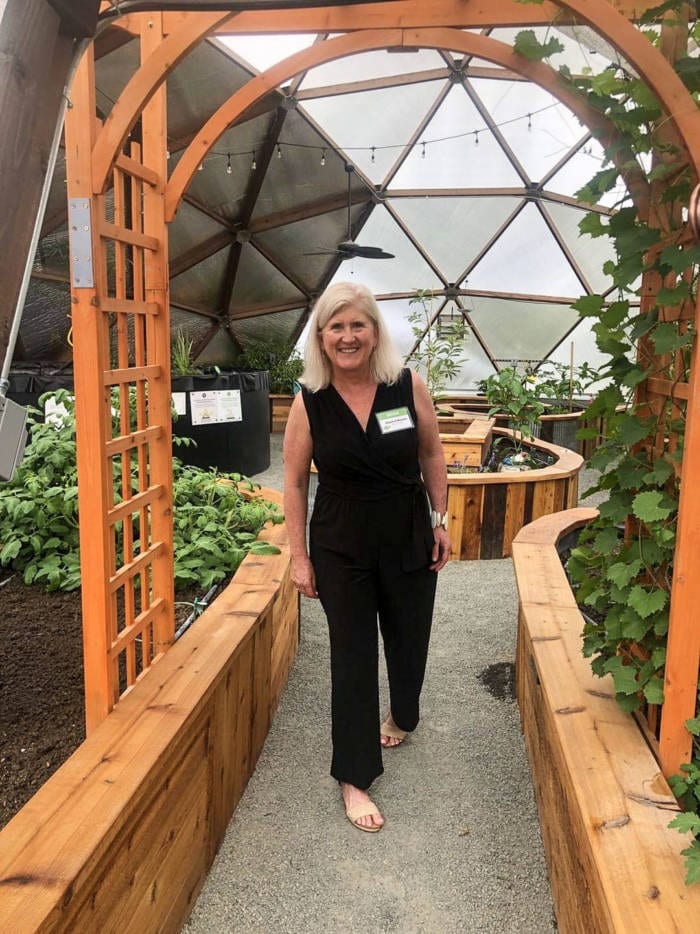 A smiling woman standing in a greenhouse, with lush raised garden beds on either side, framed by a wooden arch with a geodesic dome ceiling in the background.
