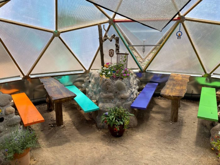 A colorful and inviting greenhouse space with vibrantly painted benches surrounding a unique rock and concrete raised garden bed centerpiece under the geometric ceiling of a geodesic dome.