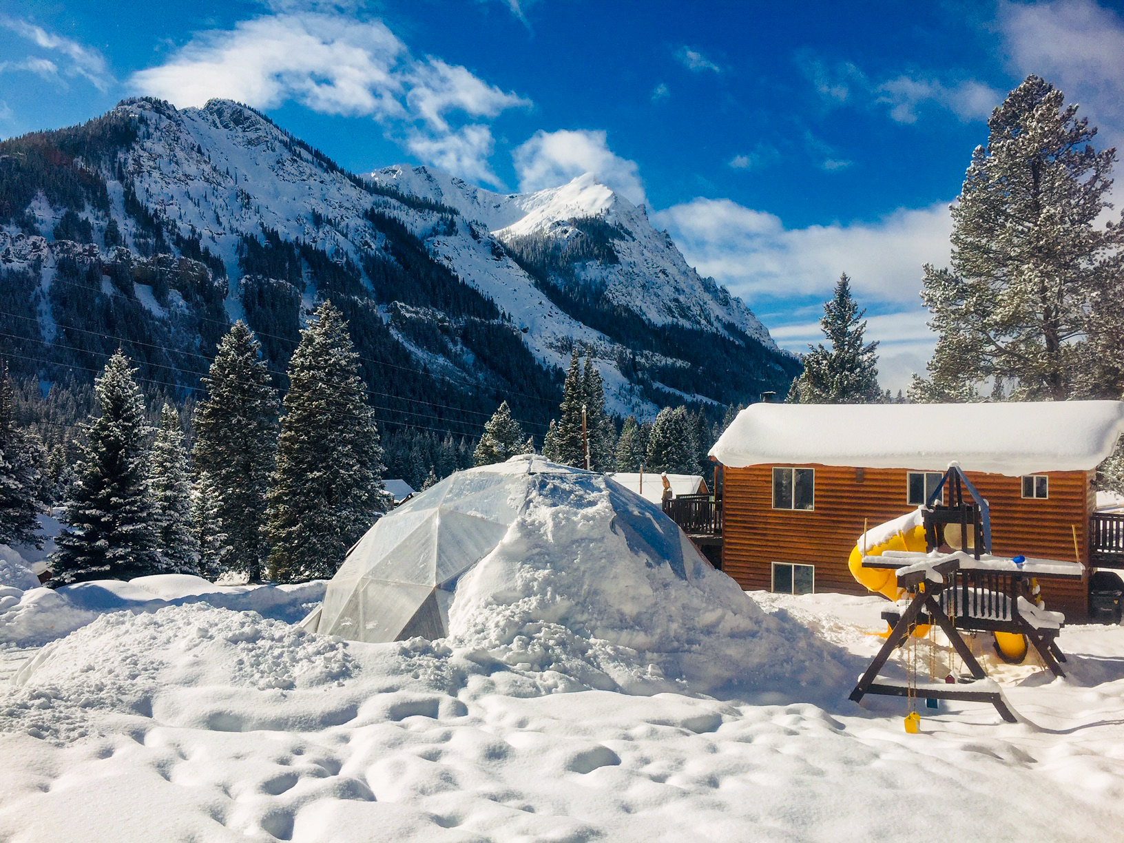 geodesic greenhouse covered in snow in the Montana mountains with a playground next to it