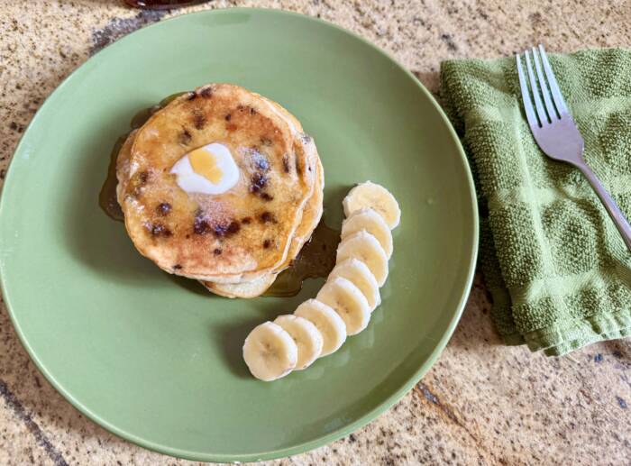Chocolate Chip Banana Pancakes on a green plate with bananas and maple syrup next to a green fork and napkin 