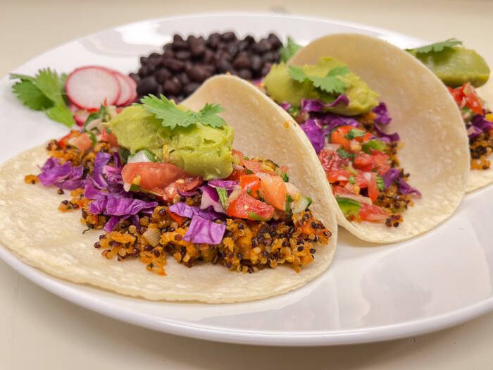Three tacos filled with vegan taco meat, topped with red cabbage, salsa, and guacamole, on a plate with black beans.